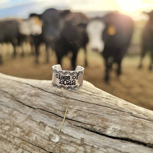 May include: A silver ring with the words "Class of 2022" engraved on it. The ring is sitting on a piece of weathered wood with a blurred background of cows.