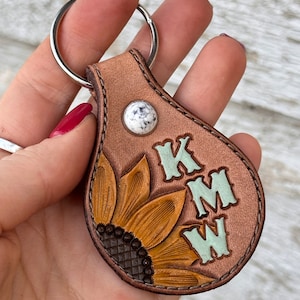 May include: A brown leather keychain with a sunflower design and the initials "KMW" in light green. The keychain has a silver ring and a white and gray stone accent.