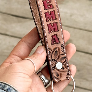 May include: A tan leather keychain strap featuring the name EMMA embossed in red capital letters. The strap is decorated with carved floral patterns, white stitching along the edges, a silver metal snap button, and a circular silver metal key ring.