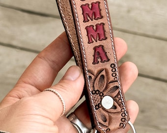 May include: A brown leather keychain with a floral design and the name "EMMA" embossed in red. The keychain has a silver key ring.