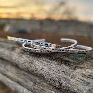 Op de afbeelding: Drie zilveren armbanden met geometrische ontwerpen. De armbanden liggen op elkaar gestapeld op een houten oppervlak.