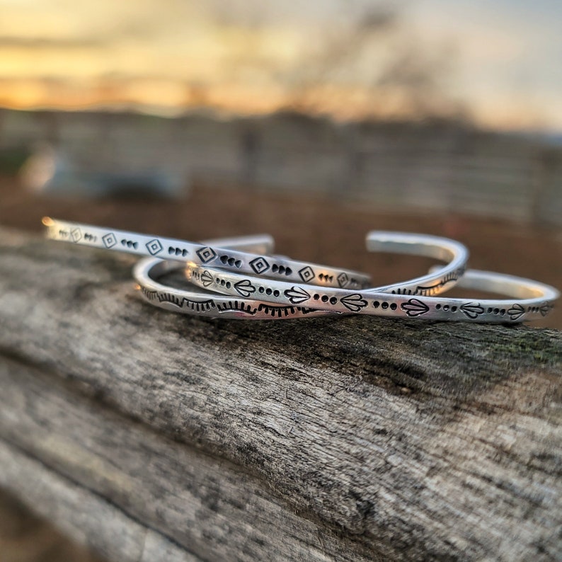 Op de afbeelding: Drie zilveren armbanden met geometrische ontwerpen. De armbanden liggen op elkaar gestapeld op een houten oppervlak.