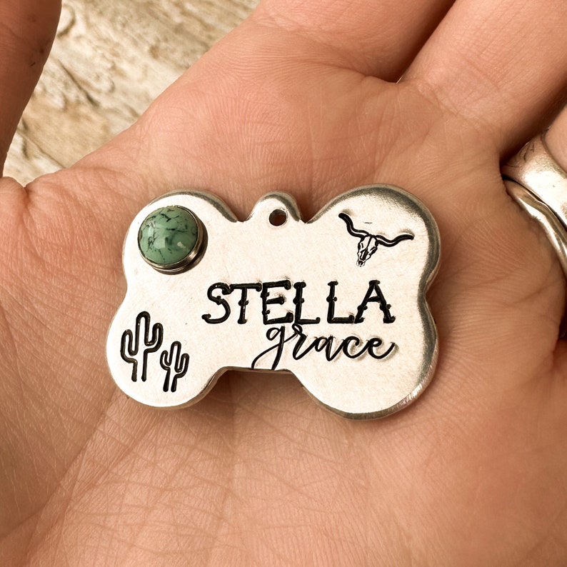 K&ouml;nnte beinhalten: Eine knochenf&ouml;rmige Haustiermarke aus silberfarbenem Metall mit der Gravur STELLA grace, einem kleinen Stiersch&auml;del-Symbol und zwei Kaktus-Symbolen. Ein runder, hellgr&uuml;ner Stein mit dunkler Maserung ist oben links in das Metall eingelassen.