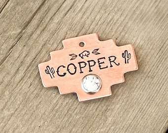 May include: A copper-colored metal tag with a southwestern design. The tag features a buffalo silhouette, arrows, and the word "COPPER" with a cactus on each side. A white stone is attached to the tag.