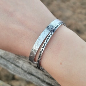 Op de afbeelding: Twee zilveren armbanden met ingewikkelde bloemmotieven. De armbanden zijn gestapeld op de pols van een persoon.