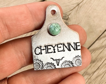 May include: Silver cow tag with the name "CHEYENNE" engraved on it. The tag is decorated with a turquoise stone and a skull with horns. The tag is surrounded by a border of sunflowers.