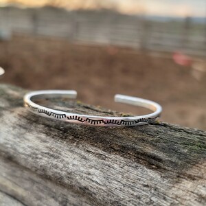 Op de afbeelding: Een zilveren armband met een herhalend patroon van kleine, gebogen lijnen.