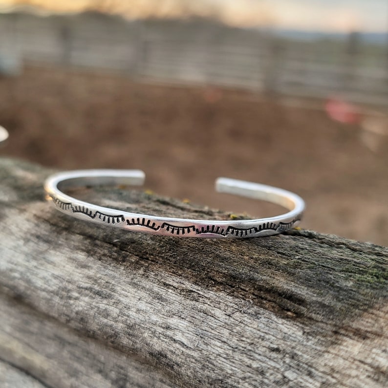 Op de afbeelding: Een zilveren armband met een herhalend patroon van kleine, gebogen lijnen.