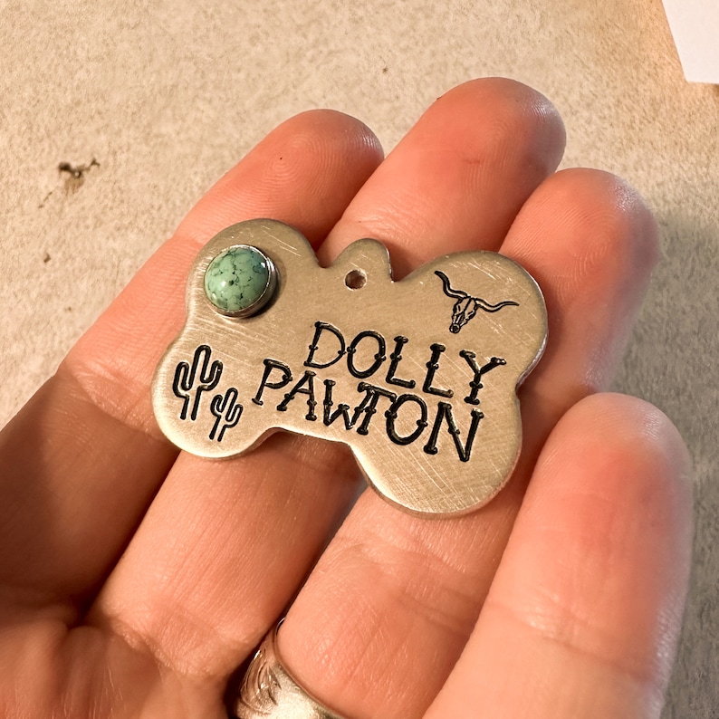 K&ouml;nnte beinhalten: Eine knochenf&ouml;rmige Haustiermarke aus Metall mit geb&uuml;rstetem Silberfinish. Die Marke zeigt den eingravierten Text DOLLY PAWTON, ein kleines Longhorn-Sch&auml;del-Symbol und zwei Kaktus-Symbole. Ein runder, gesprenkelter t&uuml;rkisfarbener Stein ist oben eingesetzt.