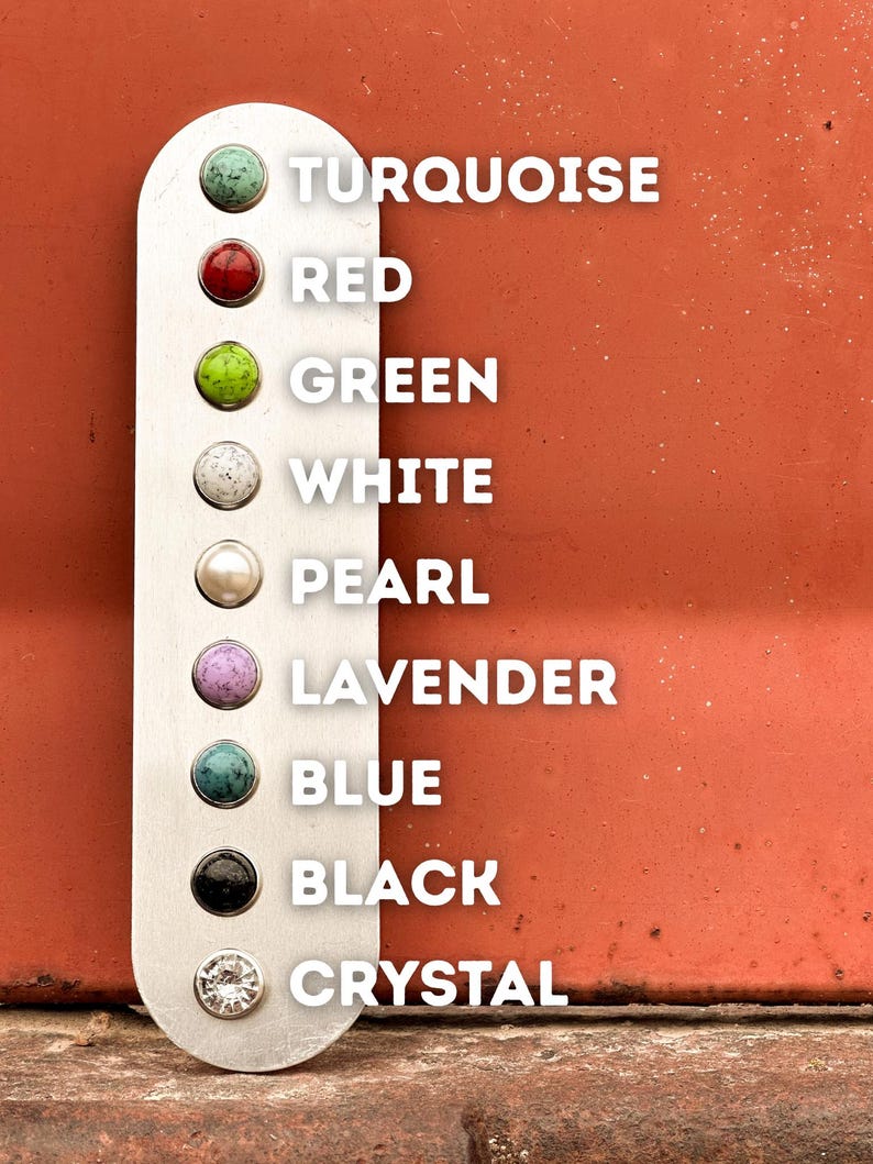 K&ouml;nnte beinhalten: Ein vertikaler, silberfarbener Metallstreifen mit neun kreisf&ouml;rmigen Einlagen, die mit wei&szlig;em Text beschriftet sind: Turquoise, Red, Green, White, Pearl, Lavender, Blue, Black und Crystal. Jede Einlage zeigt eine eigene Farbe oder Textur.