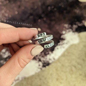 May include: Two silver rings with the words "RPS" and "ALS" engraved on them. The rings are intertwined and have small heart symbols between the letters.