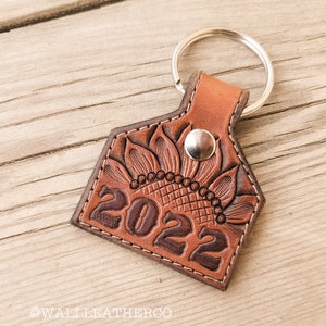 May include: A brown leather keychain with a sunflower design and the year 2022 embossed on it. The keychain has a silver metal ring and a snap closure. The edges are stitched with white thread.