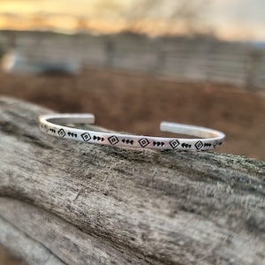 Op de afbeelding: Een zilveren armband met een gegraveerd geometrisch patroon. Het patroon bestaat uit kleine vierkanten en stippen.