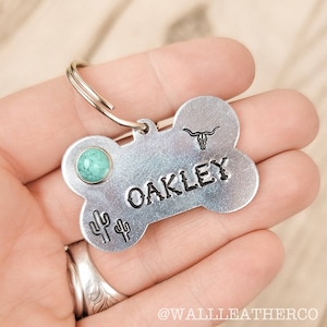 K&ouml;nnte beinhalten: Eine knochenf&ouml;rmige Haustier-ID-Marke aus Metall mit dem in Schwarz eingestanzten Namen OAKLEY, einem kleinen T&uuml;rkis-Inlay, einem eingestanzten Longhorn-Sch&auml;del-Symbol und zwei kleinen Kaktus-Symbolen. Ein silberfarbener Metallring zur Befestigung ist enthalten.