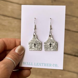 May include: A pair of silver-colored earrings with a cross design. The earrings are shaped like a tag with a floral pattern and a small cross in the center. The earrings are displayed on a white card with the text "WALL LEATHER CO."