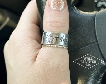 Puede incluir: Un anillo de pulgar de plata con una cruz y un diseño western. El anillo se lleva en el pulgar de una persona. El texto "WALL LEATHER CO." es visible en la esquina inferior derecha de la imagen.