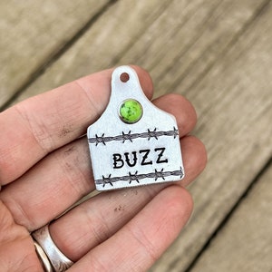 May include: A silver metal tag shaped like a cattle ear tag with a green stone in the center and the word "BUZZ" engraved on the tag. The tag is surrounded by a barbed wire design.