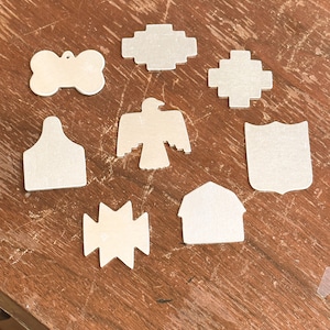 May include: A collection of eight silver-colored metal blanks in various shapes, including a bone, a cross, a bird, a shield, a barn, and geometric forms. Each piece features a smooth, flat surface with a metallic finish, arranged in a cluster.