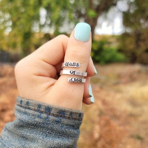 May include: A silver ring with the words "class of 2022" engraved on it. The ring is worn on a person's thumb.