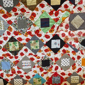 May include: A patchwork quilt with a diamond pattern. The quilt is made of various fabrics in different colours and patterns, including red, white, black, green, and blue. The quilt is decorated with a floral pattern of red poppies.