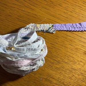 May include: A bundle of fabric scraps in various colors and patterns, including white, pink, and purple, is tied together with a purple fabric strip.