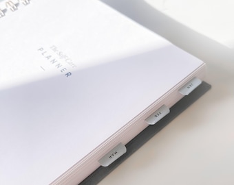May include: A white spiral-bound planner with the text "The Self-Care Planner" and month tabs labeled JAN, FEB, MAR, and APR. The planner has a gray cover and is open to a blank page.