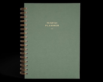 May include: A green spiral-bound planner with gold lettering that reads "The Self-Care Planner." The cover has a textured appearance. The spiral binding is gold-colored.