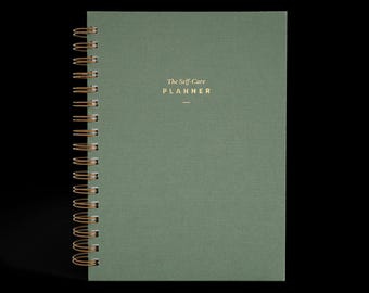 May include: A green spiral-bound planner with gold lettering that reads "The Self-Care Planner." The cover has a textured appearance. The spiral binding is gold-colored.