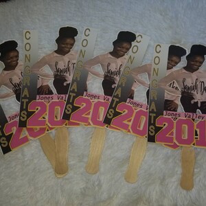 May include: Five graduation fan decorations with a photo of a young person in a pink shirt and black pants. The fans have the words "CONGRATS" and the years "2020" and "2019" printed on them.