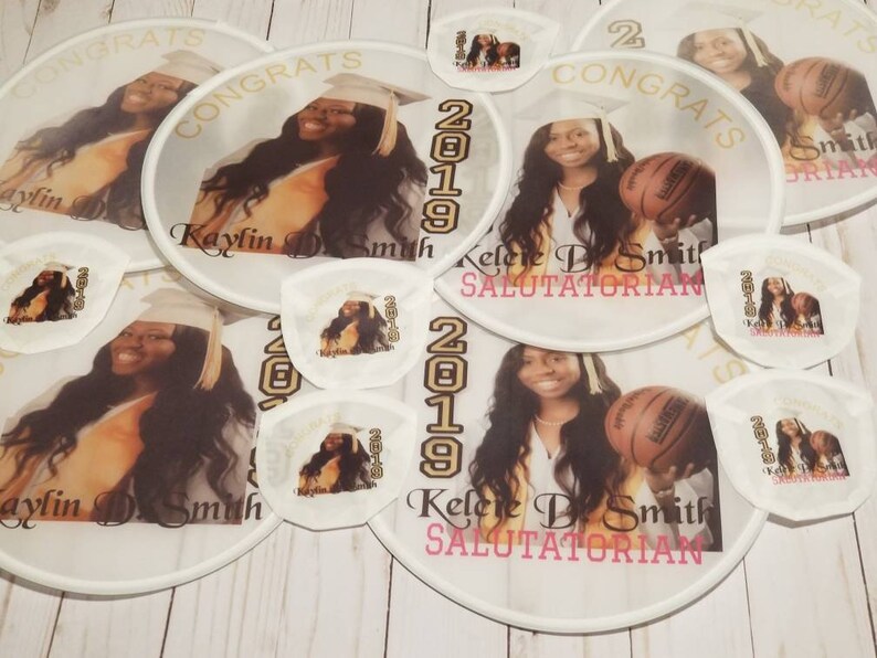 May include: A set of white hand fans with a gold trim and the text "CONGRATS" and "2019". The fans feature photos of two graduates, one with a basketball and the other with a graduation cap. The text "Kaylin D. Smith" and "Kelere D. Smith SALUTATORIAN" is also included.