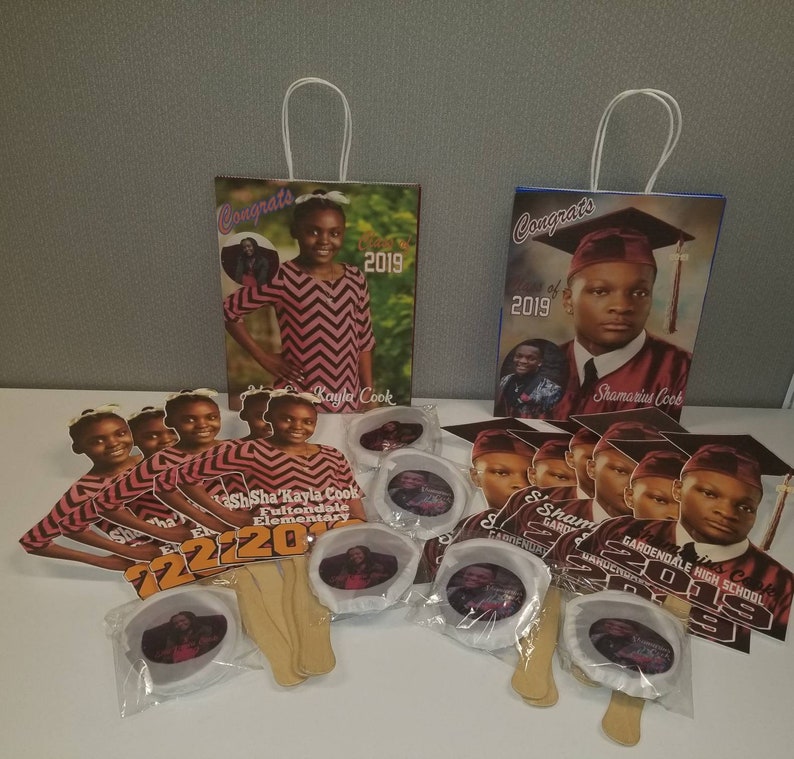May include: Graduation party favors featuring a red and black color scheme. The favors include paper fans, paper bags, and round paper favors with photos of the graduates. The text "Congrats" and "Class of 2019" are visible on the bags. The text "Sha'Sha Kayla Cook Fultondale Elementary" and "Shamarius Cook Gardendale High School" are visible on the fans.