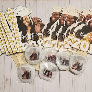 May include: A set of graduation party decorations featuring a white and gold color scheme. The decorations include a banner with the word "Salutatorian" printed in gold letters, a graduation cap cutout with a photo of a graduate, and a sign with the graduate's name and graduation year. The decorations are on a white wooden background.