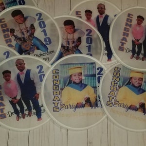 May include: A set of eight round, white, paper fans with blue and gold trim. Each fan features a photo of a child in a graduation cap and gown, along with the text "CONGRATS" and the year "2019". The fans also include the child's name and the name of the school.