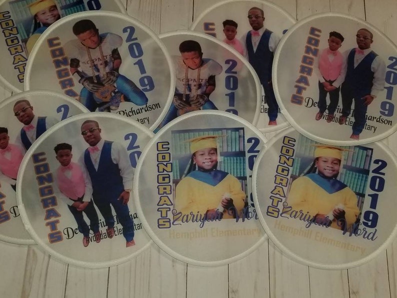 May include: A set of eight round, white, paper fans with blue and gold trim. Each fan features a photo of a child in a graduation cap and gown, along with the text "CONGRATS" and the year "2019". The fans also include the child's name and the name of the school.