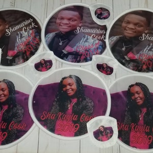 May include: Set of eight white round graduation party decorations with photos of two graduates. The decorations feature the names "Shamarius Cook" and "Sha Kayla Cook" and the year "2019".
