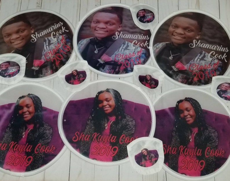 May include: Set of eight white round graduation party decorations with photos of two graduates. The decorations feature the names "Shamarius Cook" and "Sha Kayla Cook" and the year "2019".