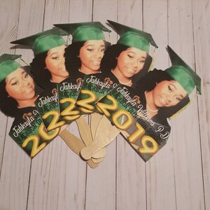 May include: A set of six graduation party fan decorations featuring a photo of a graduate wearing a green cap and gown. The fans are decorated with gold lettering that reads "Jahkayla Williams, P.D. Jackson High School 2019" and "Jahkayla Jackson 2022".