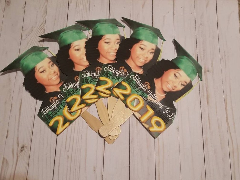 May include: A set of six graduation party fan decorations featuring a photo of a graduate wearing a green cap and gown. The fans are decorated with gold lettering that reads "Jahkayla Williams, P.D. Jackson High School 2019" and "Jahkayla Jackson 2022".