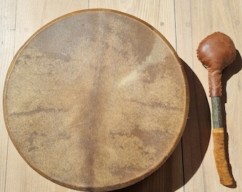 May include: A brown leather drum with a wooden frame and a brown leather beater. The drum is sitting on a wooden floor.