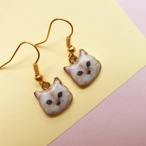 May include: A pair of gold dangle earrings with white cat face charms. The cat faces have a realistic painted design.