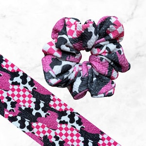 May include: A fabric scrunchie and a matching long fabric strip featuring a vibrant pattern of hot pink and white checkered squares interspersed with black and white cow print spots. The material has a textured, slightly crinkled appearance.