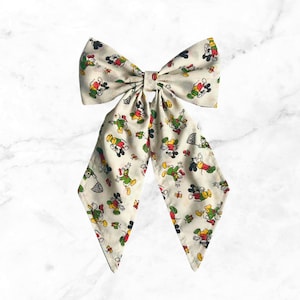 May include: A large cream-colored bow tie featuring a repeating pattern of Mickey Mouse and Donald Duck in various poses. The bow tie has long, flowing tails and is made of a lightweight fabric.