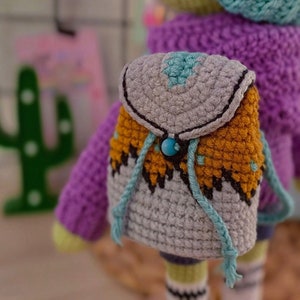 Puede incluir: Una muñeca de crochet que lleva un suéter morado y un gorro azul y blanco. La muñeca lleva una pequeña mochila de crochet gris y naranja con un botón azul.