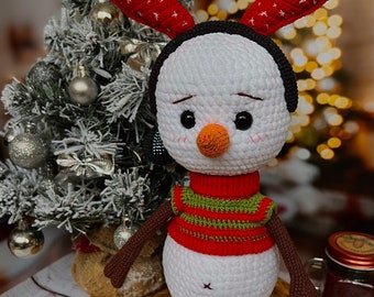 May include: A white crocheted snowman with brown arms and legs, wearing a red and green striped sweater and a black headband with red antlers. The snowman is standing on a woven wicker mat.