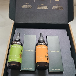 Könnte beinhalten: Eine schwarze Pappschachtel mit zwei braunen Glastropfflaschen der Marke Purdy & Figg, eine grün etikettiert als Basil & Spearmint und eine orange als Orange & Clove, inklusive zwei passender Schachteln und Anweisungen im Deckel.