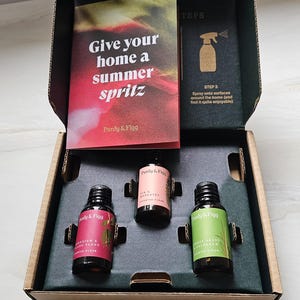 Puede incluir: Una caja de cartón que contiene tres frascos de vidrio pequeños con tapas negras y etiquetas de colores, marcados Purdy & Figg. Una tarjeta impresa dice Give your home a summer spritz, junto a una tarjeta de instrucciones con un icono de spray.