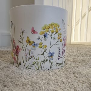 May include: A cylindrical white wastebasket featuring a colorful botanical print. The design displays various wildflowers in shades of yellow, pink, and blue, interspersed with small butterflies. The surface is smooth and matte in appearance.