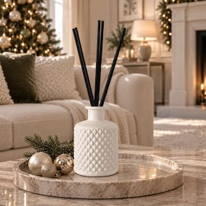 May include: A white ceramic reed diffuser bottle with a textured diamond pattern on the exterior, featuring four thin black reeds extending from the top. The bottle is placed on a round, light-coloured stone tray alongside two small metallic ornaments.