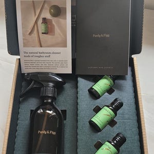 Könnte beinhalten: Eine offene Box mit Badezimmer-Reinigungsprodukten von Purdy & Figg. Das Set enthält eine Sprühflasche, kleine Flaschen, eine Broschüre und ein Waschlappen. Der Text auf der Broschüre lautet "Explore Our Scents".