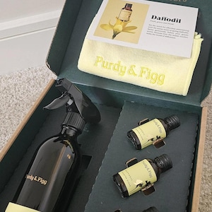 Puede incluir: Un kit de limpieza Purdy & Figg en una caja verde oscuro que contiene una botella de spray negra con base amarilla, tres botellas pequeñas de vidrio ámbar etiquetadas como Daffodil y un paño de microfibra amarillo con el logo bordado.